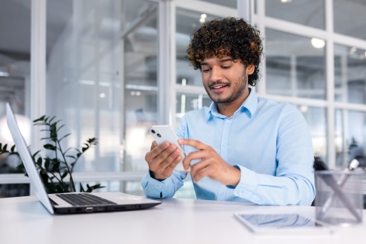 Young man using mobile phone sitting at desk with laptop